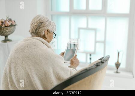 Vieille femme aux cheveux gris se détendant à la maison avec UNE tablette dans sa main. Banque D'Images