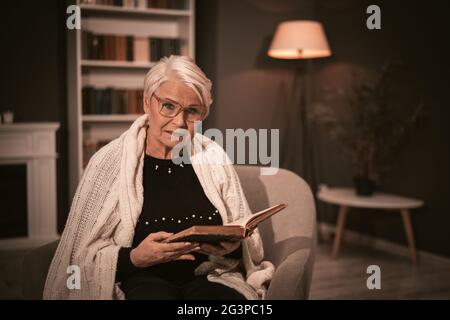 Une femme âgée lit le livre ancien assis dans une chaise. Banque D'Images