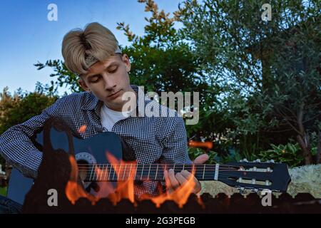 Joli homme blond avec guitare acoustique. Un jeune homme dans un bandana blanc et une chemise à carreaux est assis près du feu et joue un instrument de musique Banque D'Images