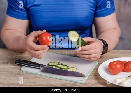 Un homme tient un concombre et une tomate dans ses mains. Il y a un couteau sur la planche, et un concombre coupé sur la table Banque D'Images