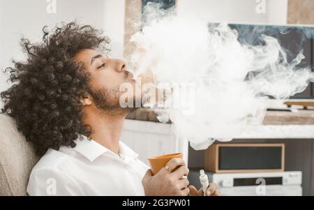 Jeune homme fume de la cigarette électronique à la maison. Le fumeur exhache un gros nuage de fumée, une vue prifile de l'arabe fume de boire Banque D'Images