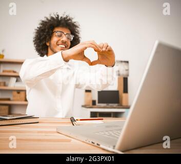 Un jeune homme passe un appel vidéo, communique avec ses amis via une liaison vidéo. Un homme arabe montre un signe de coeur avec ses mains exprimant lo Banque D'Images