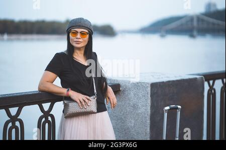 Femme voyageur en lunettes de soleil reposant sur le remblai de la rivière. Belle femme asiatique avec valise voyage dans la ville européenne. Banque D'Images