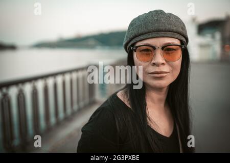 Jolie femme asiatique sur la rive de la rivière. Jolie brunette d'âge moyen en lunettes de soleil jaunes regarde l'appareil photo sur le fond de l'emba Banque D'Images