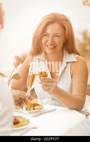 Une femme heureuse tient un verre de vin blanc souriant tout en regardant son mari. Un couple caucasien mature célèbre un anniversaire de mariage Banque D'Images