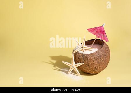 Noix de coco avec étoiles de mer, sable et parasol sur fond jaune en plein soleil, concept de voyage d'été, gros plan. Banque D'Images