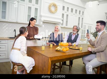 Jeune belle mère apportant un gâteau avec des bougies pour petite fille mignonne fille célébrant son anniversaire, heureux grands-parents et père assis derrière le bureau de cuisine et de filmer des moments spéciaux Banque D'Images