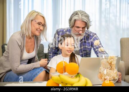 Utilisation des technologies modernes. Petite fille mignonne avec grand-père positif et grand-mère portant des lunettes regardant l'écran d'ordinateur portable regarder des vidéos assis sur un canapé confortable dans le salon à la maison Banque D'Images