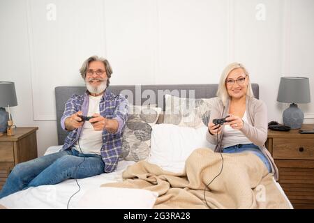 Grand-père et grand-mère excités jouant aux jeux vidéo tout en étant confortablement assis sur le lit tenant les manettes et en appréciant le temps de loisirs ensemble à la maison, couple senior mari et femme s'amusant Banque D'Images