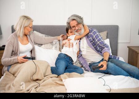 Petite fille adorable petite-fille embrassant grand-père souriant lorsqu'il joue à des jeux vidéo avec grand-mère tout en étant assis sur le lit. Bonne famille passant du temps ensemble le week-end à la maison Banque D'Images