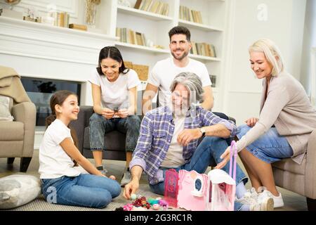 Grande famille heureuse multigénérationnelle jouant avec un enfant mignon dans le salon à la maison. Petite fille montrant sa nouvelle maison de poupée et ses jouets à son père, sa mère et ses grands-parents, assis sur le sol et en riant Banque D'Images