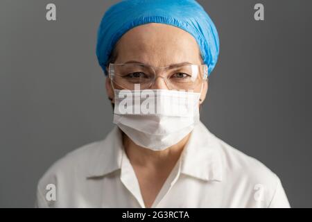 Visage de médecin féminin, gros plan. Une femme mature dans un masque de protection et des lunettes de protection regarde la caméra. Isolé sur gris Banque D'Images
