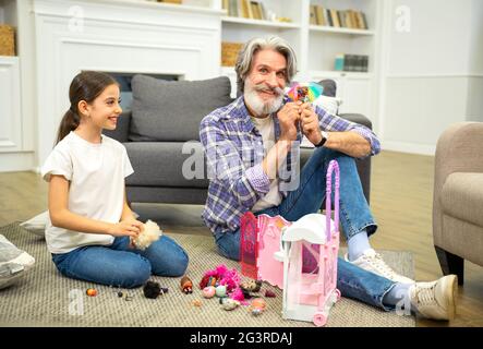 Petite fille souriante petite-fille jouant avec grand-père tout en passant un week-end chez les grands-parents, montrant maison de poupée à grand-père tout en étant assis dans le salon au sol et en appréciant le temps ensemble Banque D'Images