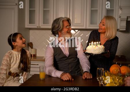 Une grand-mère heureuse apporte un gâteau aux bougies à son mari souriant qui fête son anniversaire avec sa famille. La petite petite fille mignonne est excitée d'attendre que la grand-père souffle des bougies Banque D'Images