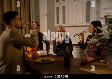 Dîner de vacances. Jeunes couples et grands-parents buvant du champagne tout en célébrant l'anniversaire de grand-père dans la cuisine à la maison portant une tenue de fête formelle. Concept de célébration familiale Banque D'Images