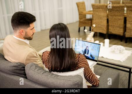 Une jeune femme et un mari parlent de problèmes avec un psychologue de famille par appel vidéo sur un ordinateur portable tout en étant assis sur un canapé à la maison, vue arrière. Jeune couple ayant consultation en ligne avec le médecin Banque D'Images