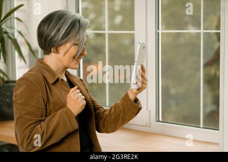 Une femme assez mûre communique en ligne sur une tablette numérique située près de la fenêtre. Femme souriante en contact avec des amis par internée Banque D'Images