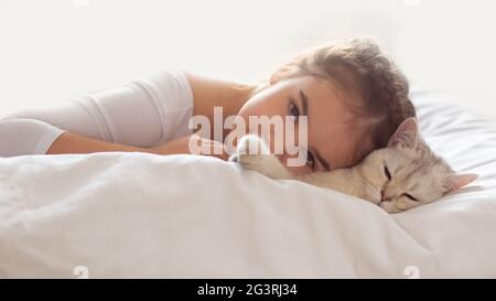 Bannière large. Une belle petite fille en vêtements blancs, se trouve sur un lit propre et blanc avec un chat britannique blanc. Banque D'Images