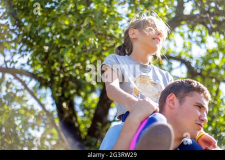 Frère défoqué de la sœur de circonscription à l'arrière. Portrait d'une fille heureuse sur les épaules de l'homme, porcgyback. Petite mouche. Famille jouant à l'extérieur. Flou d'arbre vert Banque D'Images