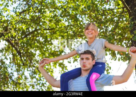 Frère défoqué de la sœur de circonscription à l'arrière. Portrait d'une fille heureuse sur les épaules de l'homme, porcgyback. Petite mouche, levez la main. Famille jouant à l'extérieur. Arbre vert Banque D'Images