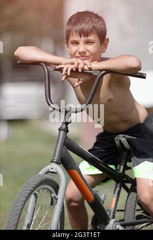 Un enfant avec un vieux vélo dans la rue. Un garçon blanc est assis sur un vélo dépassé le jour de l'été. Banque D'Images