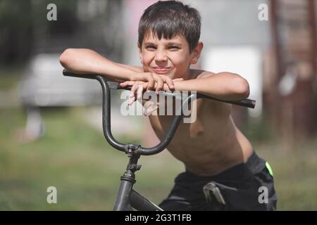 Un enfant avec un vieux vélo dans la rue. Un garçon blanc est assis sur un vélo dépassé le jour de l'été. Banque D'Images