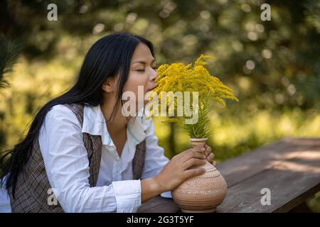 La femme de beauté sniffs l'arôme des fleurs jaunes le jour chaud dans le jardin. Brunette asiatique reposant sur une table en bois avec bouquet de Solida Banque D'Images