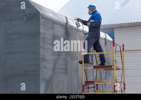 Travail industriel. Amorçage des produits métalliques à partir du pistolet du compresseur. Un travailleur en combinaison et un respirateur peint le corps d'un t Banque D'Images