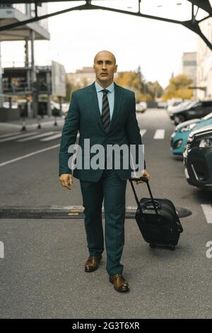 L'homme d'affaires va de l'avant en tenant sa valise en main. Un bel homme du Caucase qui passe devant les voitures garées de l'hôtel. Concept de voyage d'affaires Banque D'Images
