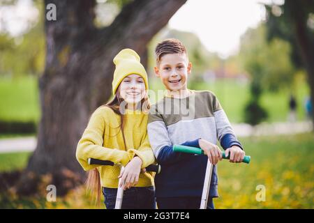 Les enfants caucasiens les jumeaux, garçons et filles, enfants d'âge scolaire, se posent ensemble par temps ensoleillé dans le parc d'automne après avoir pris le kick scoo Banque D'Images