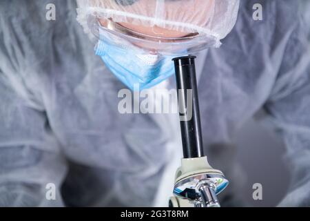 Professionnel de la santé en costume de protection blanc regardant à travers le microscope en laboratoire chimique. Rechercher le coronavirus du vaccin. COV Banque D'Images