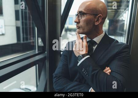 Homme d'affaires attentionné en costume avec lunettes de vue donnant sur la fenêtre ou ascenseur en verre avec vue sur la rue animée. Busi Banque D'Images