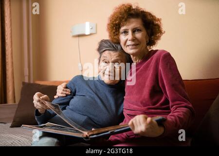 Femme âgée et sa fille adulte regardant un album photo ensemble sur un canapé dans le salon, parlant joyeusement de souvenirs. Banque D'Images