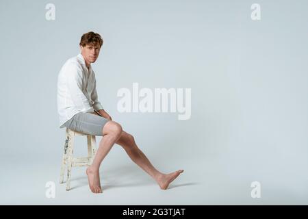 Prise de vue en longueur d'un jeune beau jeune homme assis sur une chaise portant une chemise blanche et un short gris avec une jambe en regardant CA Banque D'Images