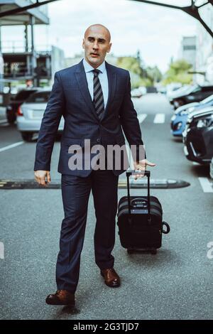 Homme d'affaires en costume sur un parking transportant une valise à pied dans les voitures garées. Homme d'affaires en costume et valise pour affaires Banque D'Images