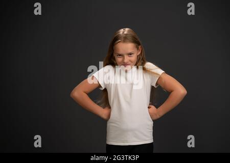 Fille bossy avec ses mains sur le côté regarder l'appareil photo porter un t-shirt blanc isolé sur du gris foncé ou du noir arrière-plan Banque D'Images