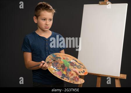 Artiste petit garçon en t-shirt bleu à l'air triste tenant une palette de peintures séchées debout à côté d'une toile sur le chevalet. PPB Banque D'Images