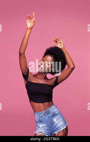 Dansant avec les mains vers le haut magnifique jeune fille afro-américaine avec des cheveux afro posant vêtu de haut noir et jeans en denim sur sale p Banque D'Images