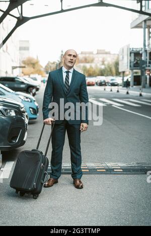 En attendant son chauffeur ou son taxi, un homme d'affaires charmant se tient sur le parking avec sa valise. Homme d'affaires en costume et valise Banque D'Images