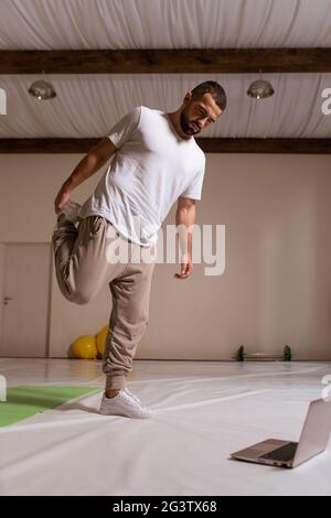 Homme de sport en t-shirt blanc et pantalon de survêtement se morçant les jambes, se réchauffant avant l'entraînement, vue latérale gros plan portrait. Stang Banque D'Images