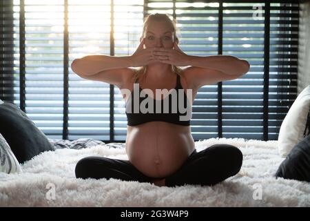 Jolie femme enceinte dans des leggins noirs et soutien-gorge couvrant la bouche avec les mains tout en étant assise sur son lit à la maison Banque D'Images