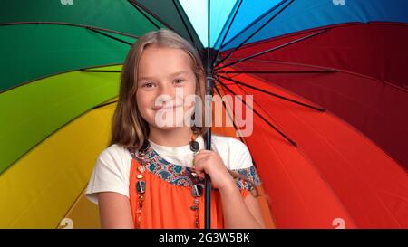 Petite fille avec parapluie de couleur arc-en-ciel dans ses mains portant la robe orange debout heureux sourire en studio tout en regardant le Banque D'Images