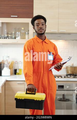 Portrait d'un sérieux jeune plombier afro-américain barbu dans des vêtements de travail orange tenant le presse-papiers et la boîte à outils tout en se tenant debout dans la cuisine Banque D'Images