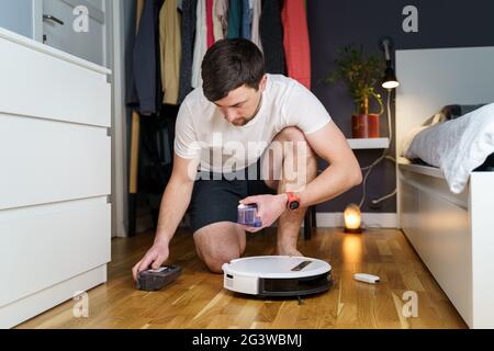 Un jeune homme de race blanche effectue la maintenance d'un robot-aspirateur blanc. Aspirateur moderne sans fil autonome. Homme réparant le robo Banque D'Images