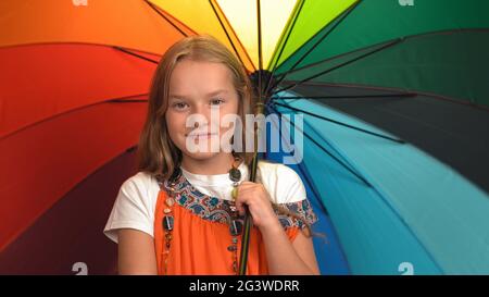 Charmante petite fille en robe orange tient un parapluie de couleurs arc-en-ciel debout et applaudissent en studio tout en regardant la caméra Banque D'Images