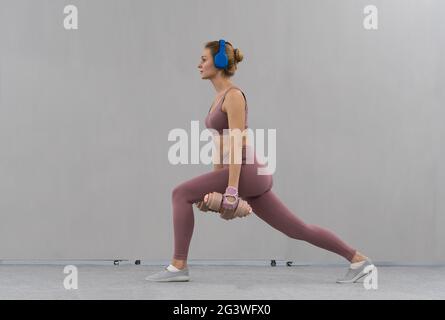 Étirez en utilisant des haltères avec les mains vers le bas jeune fille perdant du poids à la maison portant la tenue rose tout en écoutant de la musique dans bleu wirel Banque D'Images