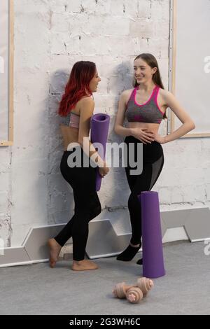 De belles jeunes femmes en sport dehors s'adapte à parler debout à côté du mur tenant un tapis de yoga, souriant les uns aux autres, debout o Banque D'Images