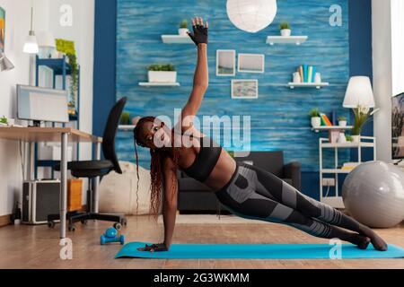 Faire des exercices physiques noirs debout dans la planche latérale sur le tapis de yoga dans la salle de séjour à la maison, faire des exercices forts, l'entraînement pour un style de vie sain. La longueur totale de l'afro-américain qui s'y emploie. Banque D'Images