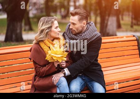 Couple amoureux heureux embrassant assis sur le banc romantique embrassé dans le parc portant des manteaux et des foulards collectant un bouquet de l'automne Banque D'Images