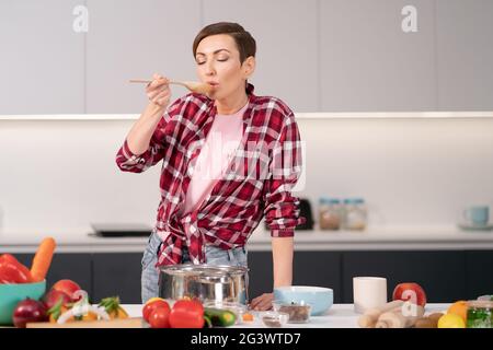 Femme essayant un plat cuit à l'aide d'une longue cuillère en bois. Femme avec un cheveux court de cuisine un dîner pour la famille debout dans le moderne Banque D'Images